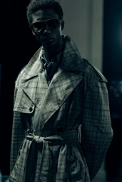 Model wearing a plaid trench coat and dark sunglasses in a moody backstage portrait for Burberry