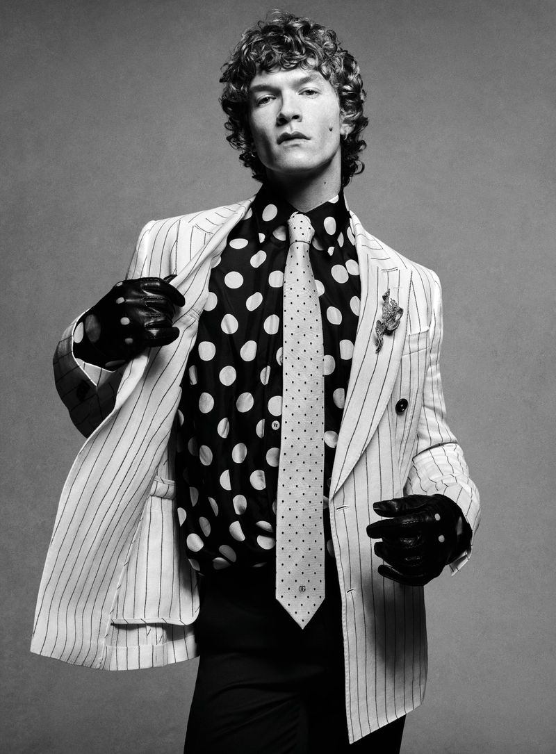 Stylish man in a pinstripe suit, polka dot shirt, and polka dot tie poses against a grey background in a fashion portrait.