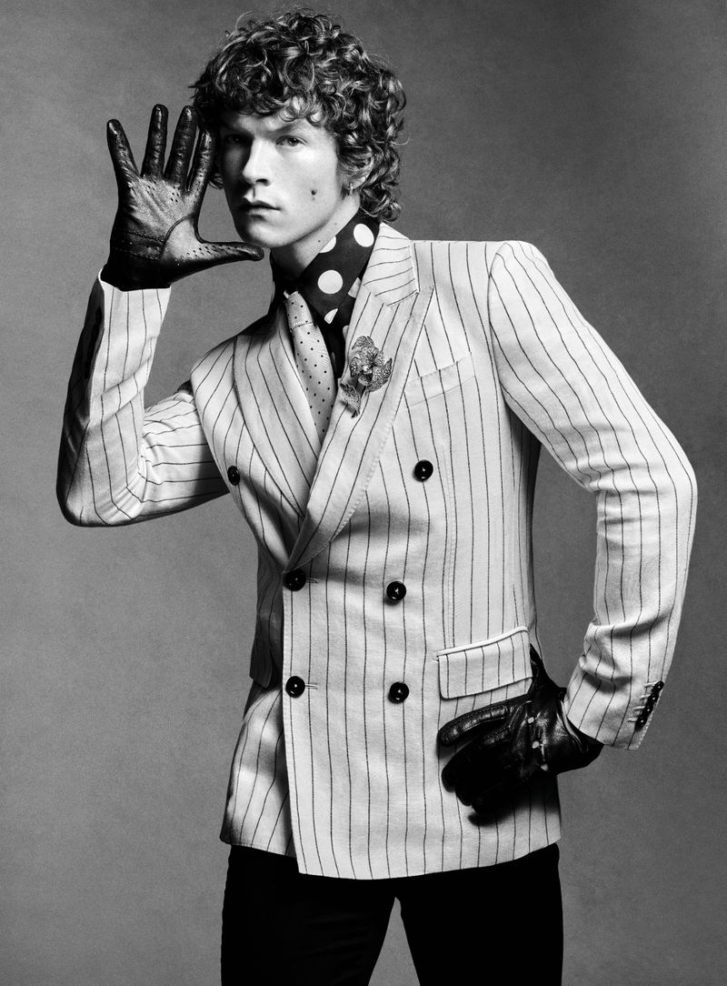 Black and white portrait of a man with curly hair wearing a double-breasted pinstripe suit, gloves, and polka dot tie, posing in a studio.