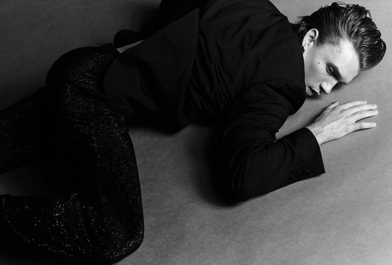 Black and white portrait of a male fashion model in a black jacket and shiny pants, lying on a studio floor in a dramatic pose.