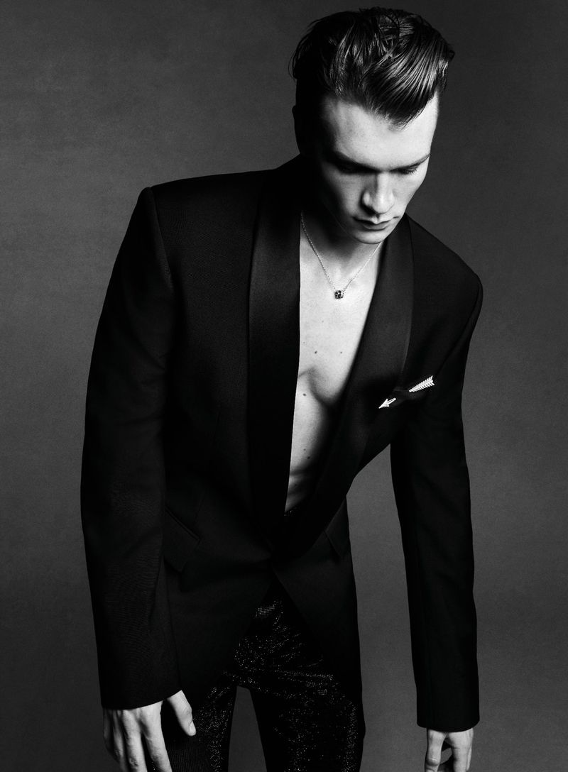 Black and white portrait of a male fashion model in a suit jacket, looking down in a photo studio setting with a gray backdrop.