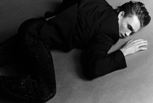 Actor Connor Storrie lying down wearing a dark jacket and trousers against a textured grey studio backdrop