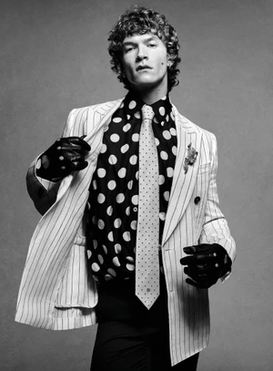 Portrait of actor Connor Storrie wearing a pinstripe jacket, polka dot shirt, tie, and leather gloves against a grey studio backdrop.