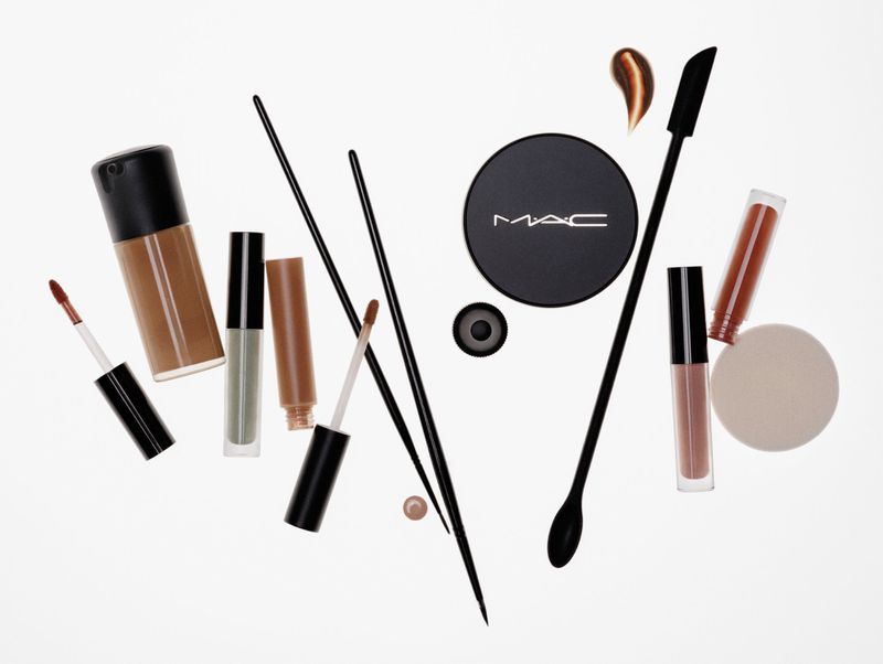 Overhead shot of makeup products including foundation, concealer, powder, lipstick, and brushes arranged on a white background.