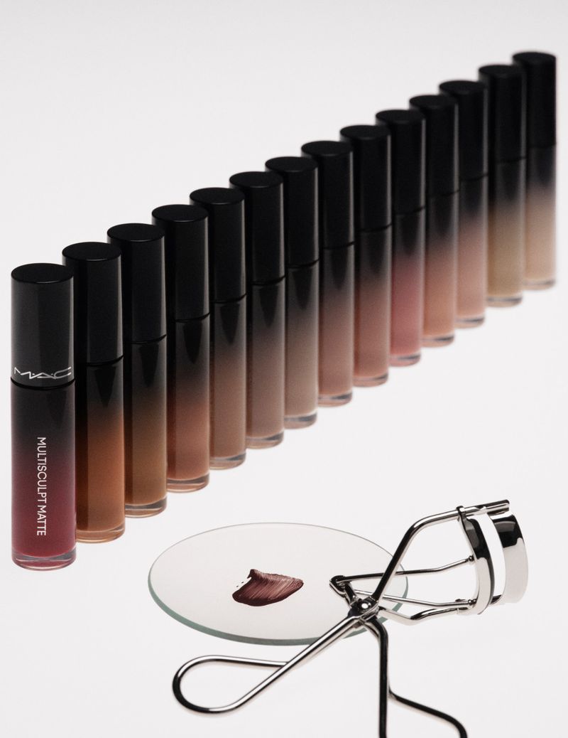 Close-up of various shades of MAC lip gloss arranged in a row, with an eyelash curler and mirrored surface on a white background.