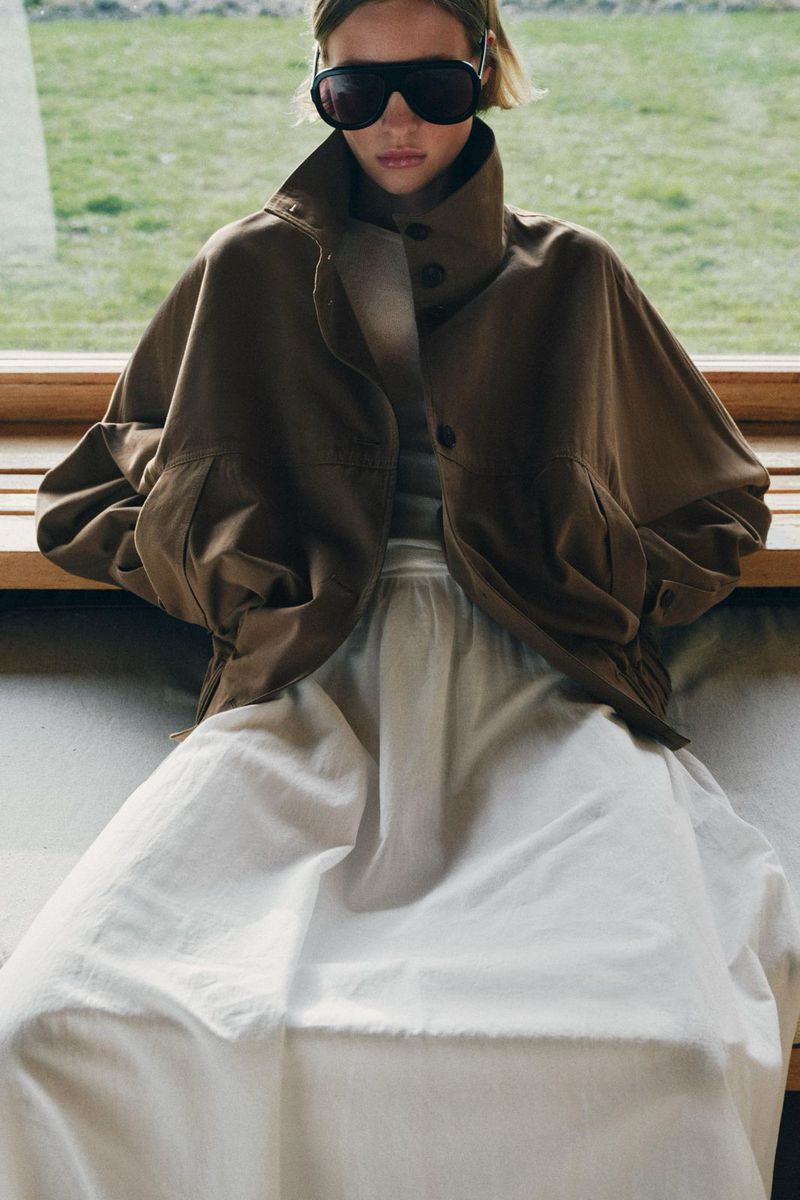 Model in oversized brown jacket and white skirt wearing dark sunglasses indoors against a window backdrop.