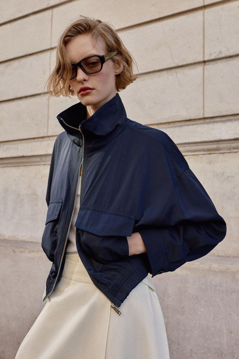 Fashion portrait of a blonde model wearing a navy jacket and cream skirt posing against a wall, accessorized with stylish sunglasses.