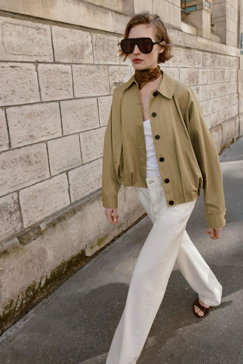 Adult female in aviator sunglasses, tan jacket, white jeans walks along urban sidewalk with brick wall. Fashion editorial outdoor shot.