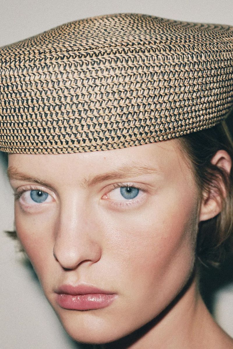 Close-up of a fair-skinned woman with bright blue eyes, wearing a beige and black woven beret, capturing a minimalist, fashion-forward aesthetic.