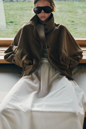 Model in oversized brown jacket and white skirt wearing dark sunglasses indoors against a window backdrop.