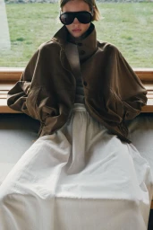Model in oversized brown jacket and white skirt wearing dark sunglasses indoors against a window backdrop.