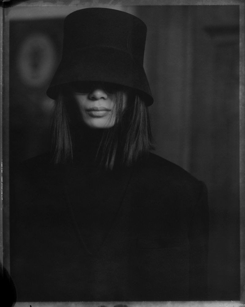 Portrait of adult female with black hair, wearing black clothing and a black bucket hat that partially obscures her face in a photo studio.