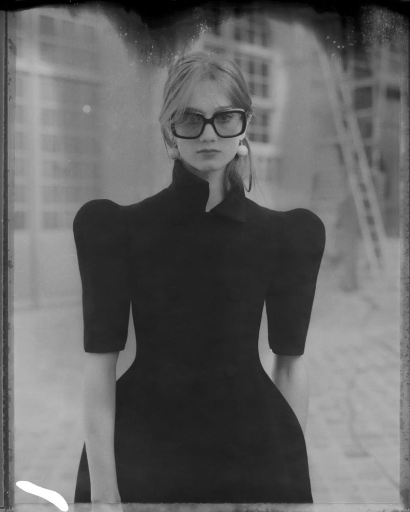 A black-and-white studio portrait of a slim woman with blonde hair wearing eyeglasses and a black dress, set against a blurred interior background.