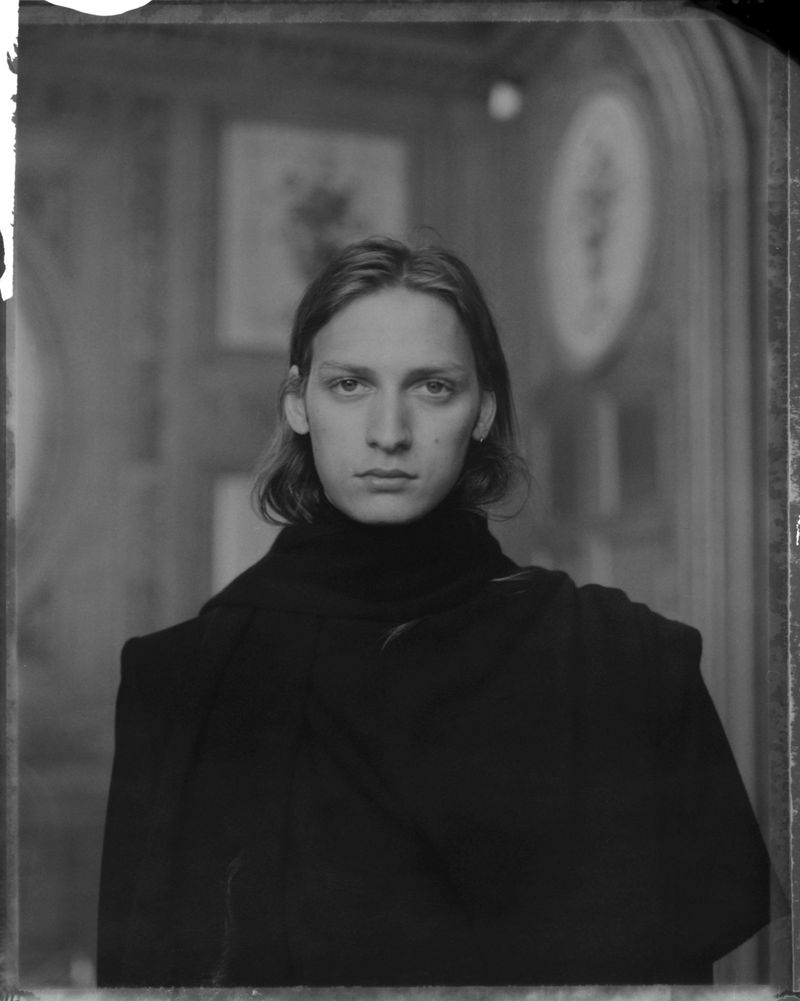 Monochrome portrait of a young adult with long hair, wearing a black turtleneck sweater, against a background of paneled walls and artwork.