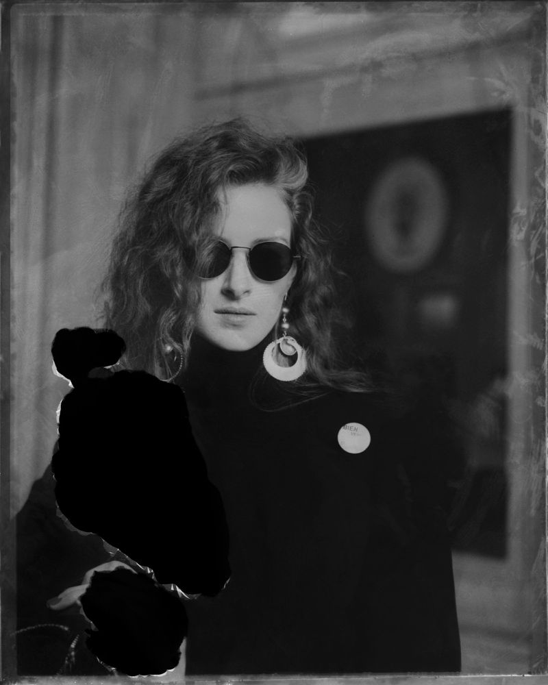 Monochrome portrait of a woman wearing sunglasses, dangling earrings, and a turtleneck sweater, with curly hair.