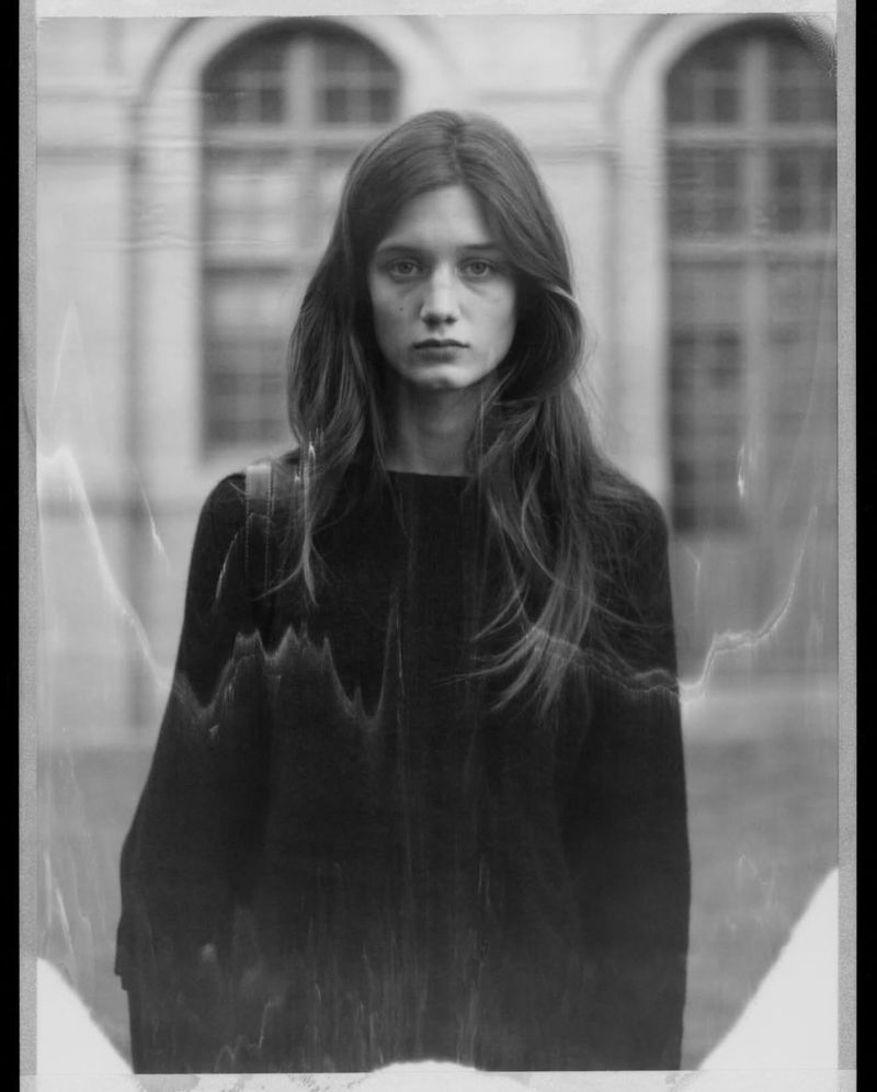 Portrait of a female model with long hair wearing a black top against a blurred architectural building exterior.