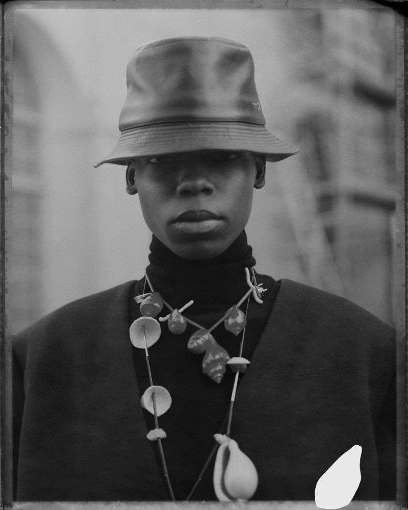Portrait of a Black adult wearing a black turtleneck, statement seashell necklace, and bucket hat, set against a neutral background.