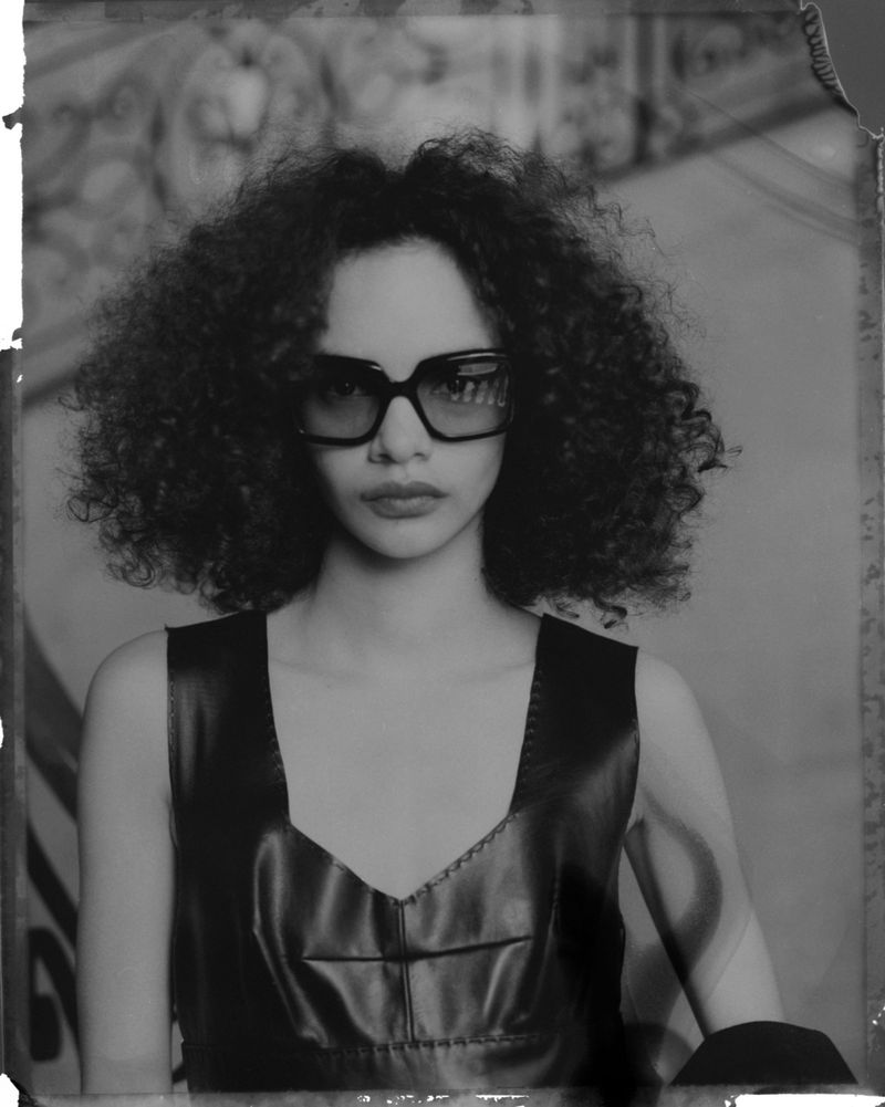 Black and white portrait of a woman with curly hair wearing sunglasses and a black top, posing against an ornate background.