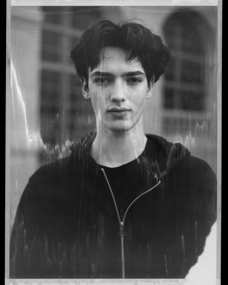 Black and white portrait of a young adult man with dark hair wearing a hooded sweatshirt, set against a blurred architectural backdrop.