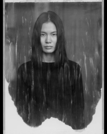 Portrait of a model with long dark hair wearing a black top against a textured monochrome backdrop.
