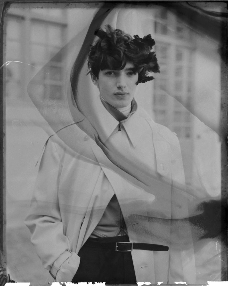 Monochrome fashion portrait of a young male model wearing a trench coat and dark pants, with a structural floral hairpiece.