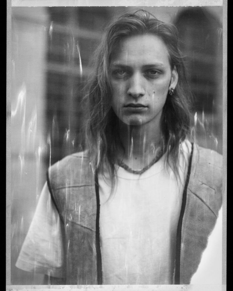 Monochrome portrait of a young adult with long hair, wearing a vest over a white tee shirt, with a blurred background.