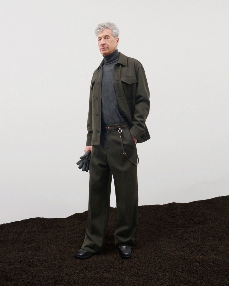 Mature man in a green collared shirt and grey turtleneck sweater, standing on dark brown soil against a light background.