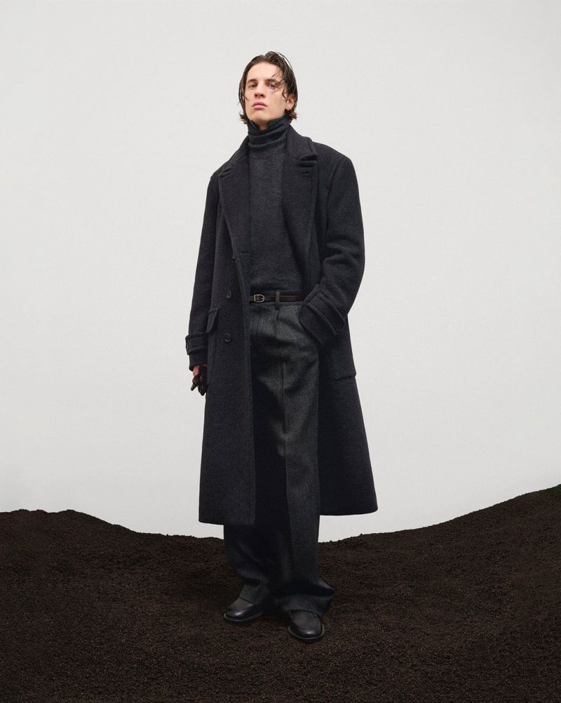A person in a grey turtleneck, trousers, and overcoat stands on dark, granular ground against a light grey background in a fashion shot.