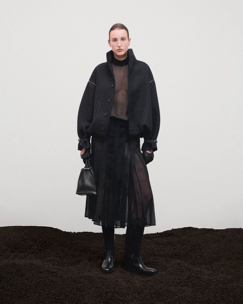 A woman in a black jacket, skirt, and boots holds a purse against a white backdrop, standing on dark brown textured surface.