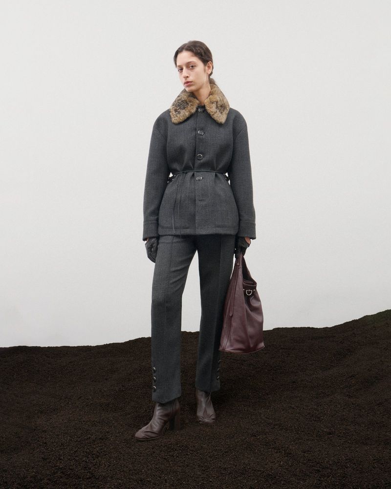 Fashion shot of a woman in a grey suit with a fur collar and boots, holding a brown leather bag, standing on dark soil against white.