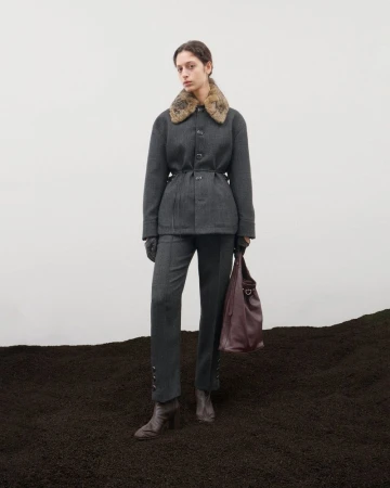 Fashion shot of a woman in a grey suit with a fur collar and boots, holding a brown leather bag, standing on dark soil against white.