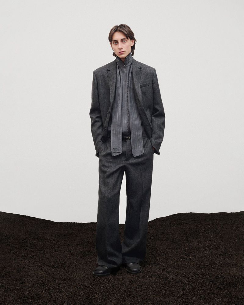 Man in grey suit, patterned scarf standing against white backdrop with textured ground. Fashion editorial-style menswear portrait.