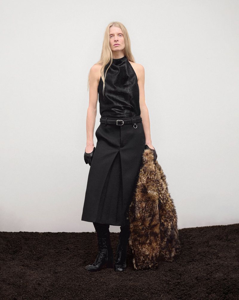 A woman with long blonde hair wears a dark halter top and midi skirt, standing on a dark surface in front of a white background.