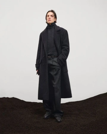 Man wearing a dark wool coat and turtleneck standing before a white backdrop above dark soil.