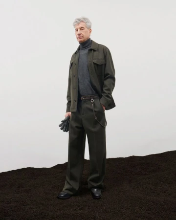 An older man standing on dark soil wearing a green jacket, grey turtleneck, and dark trousers against a white backdrop.