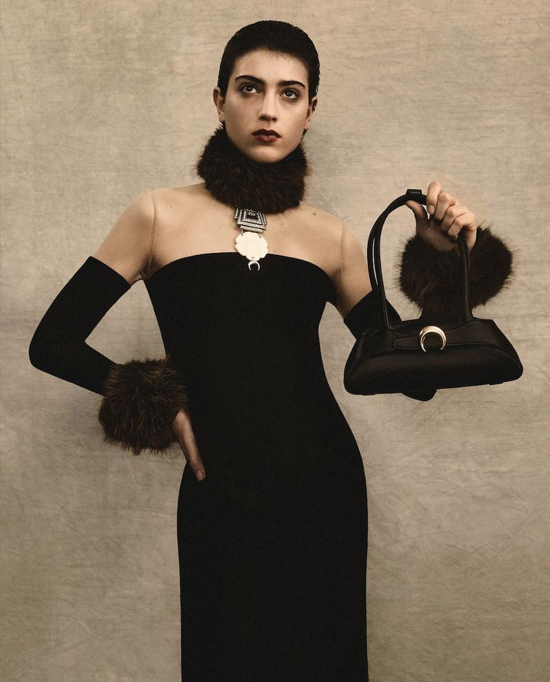 Woman in a black dress with fur details poses indoors, holding a handbag. The portrait features muted tones and simple background.