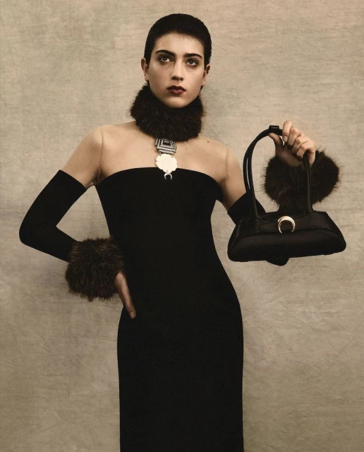 Woman in a black dress with fur details poses indoors, holding a handbag. The portrait features muted tones and simple background.