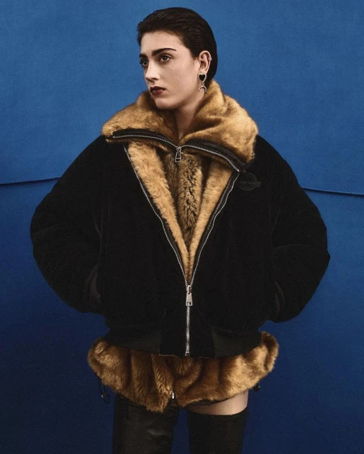 A woman poses in a black bomber jacket with fur trim against a blue backdrop, showcasing a fashion-forward and stylish look.