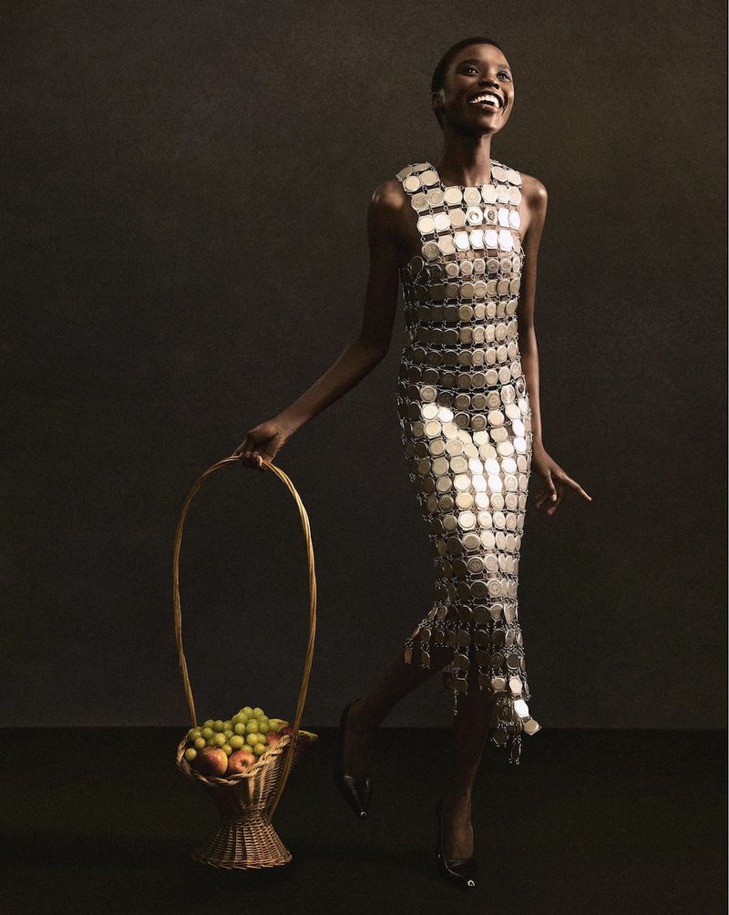 Smiling African woman in coin dress holds fruit basket in studio portrait. Metallic fashion, dark background, healthy food theme, high fashion.