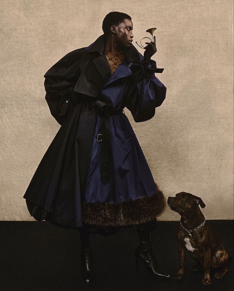 Elegant Black woman poses in a two-tone coat with a small brass horn and a dog sitting near her on a black surface.
