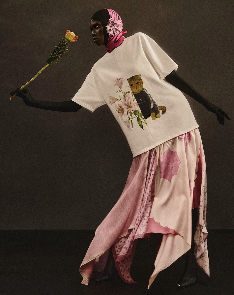 Woman in an artistic fashion pose wearing a T-shirt with a cat graphic, a pink patterned skirt, and a headscarf, holding a rose.