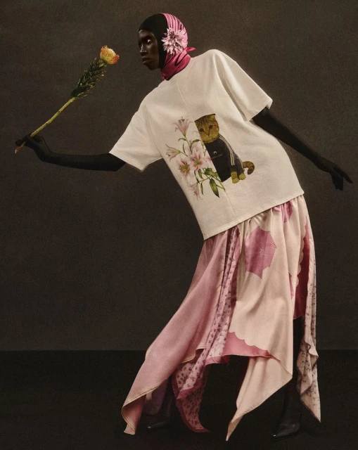 Woman in an artistic fashion pose wearing a T-shirt with a cat graphic, a pink patterned skirt, and a headscarf, holding a rose.