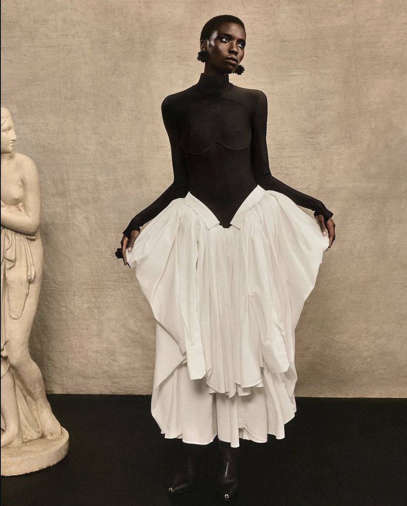 An African American woman is posing in a full-length shot, wearing a black top and white skirt, standing next to a statue indoors.