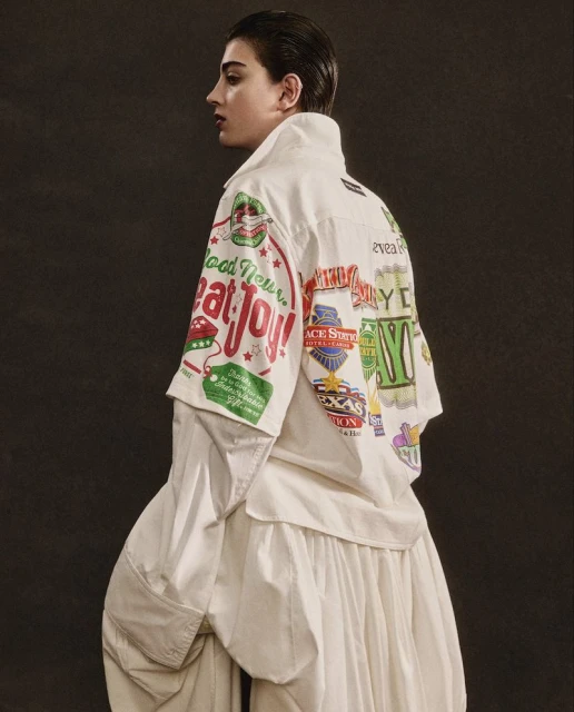 Woman in profile wearing white layered outfit with colorful graphic print jacket against a dark background in a fashion setting.