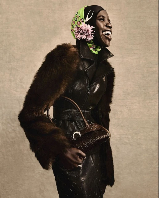 Stylish African woman in a leather outfit with fur trim, accessorized with a headscarf, jewelry, and a brown purse, smiling against a beige background.