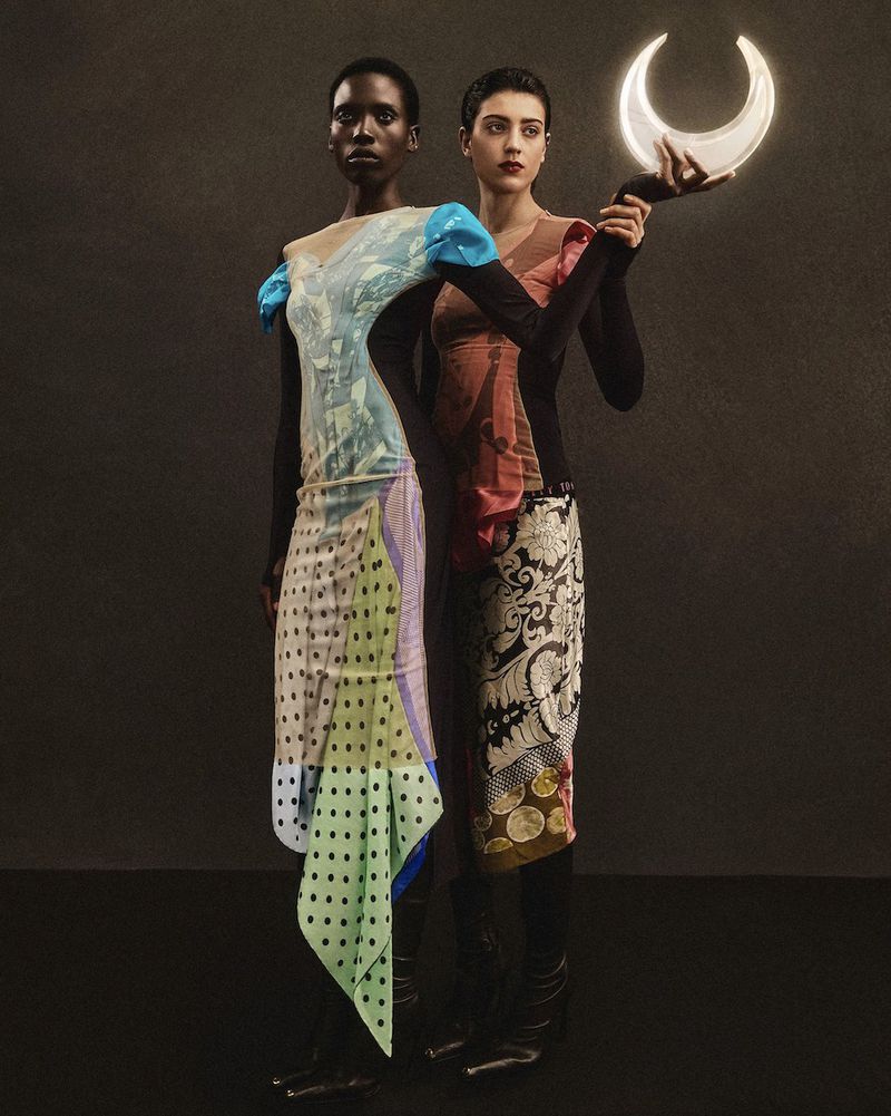 Two women stand together in a studio setting, modeling colorful dresses with distinct patterns, one holding a crescent moon prop.