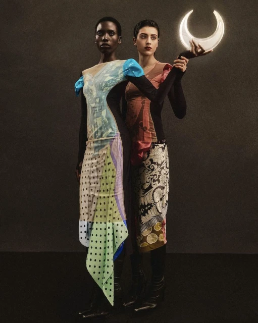 Two women stand together in a studio setting, modeling colorful dresses with distinct patterns, one holding a crescent moon prop.