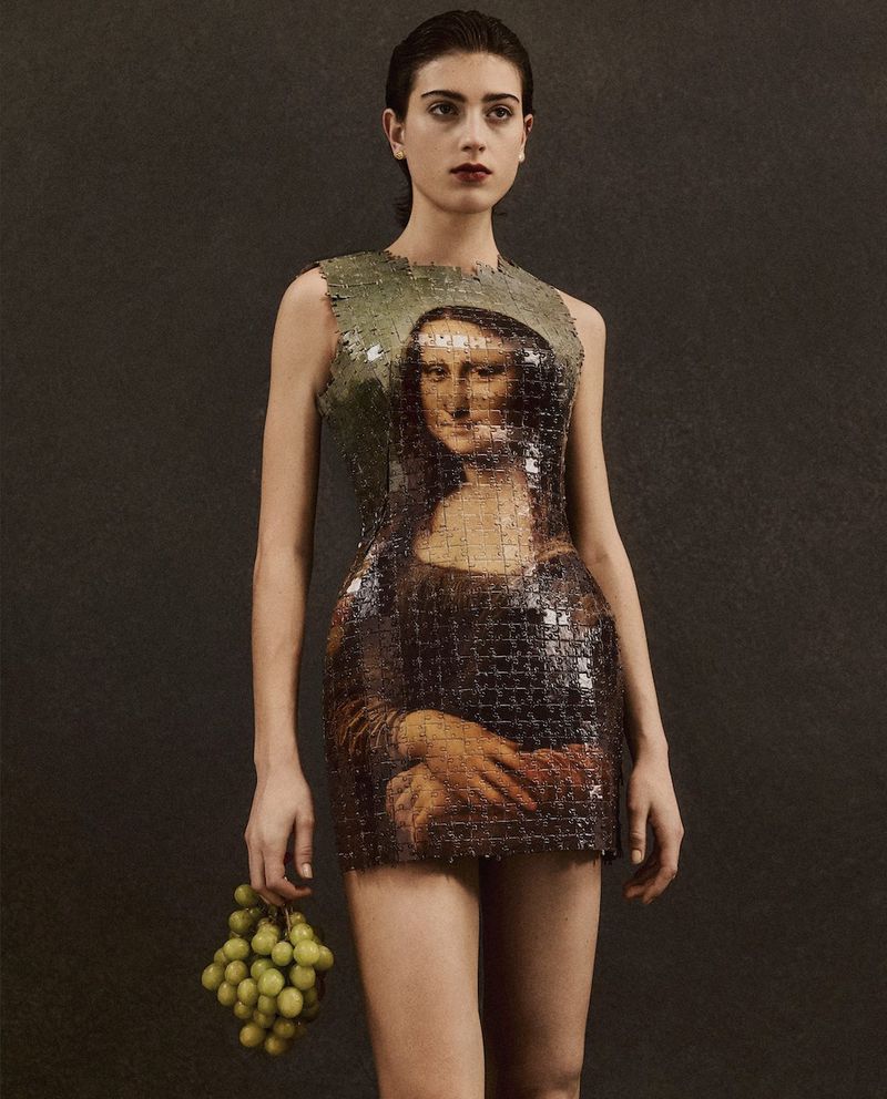 Woman in Mona Lisa dress holding grapes poses against dark backdrop in fashion shot, showcasing style and artistry with fruit prop.