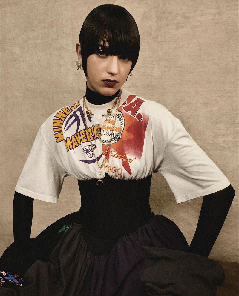Woman with bowl cut in graphic t-shirt and black corset poses against textured backdrop, accessorized with gold jewelry and earrings.