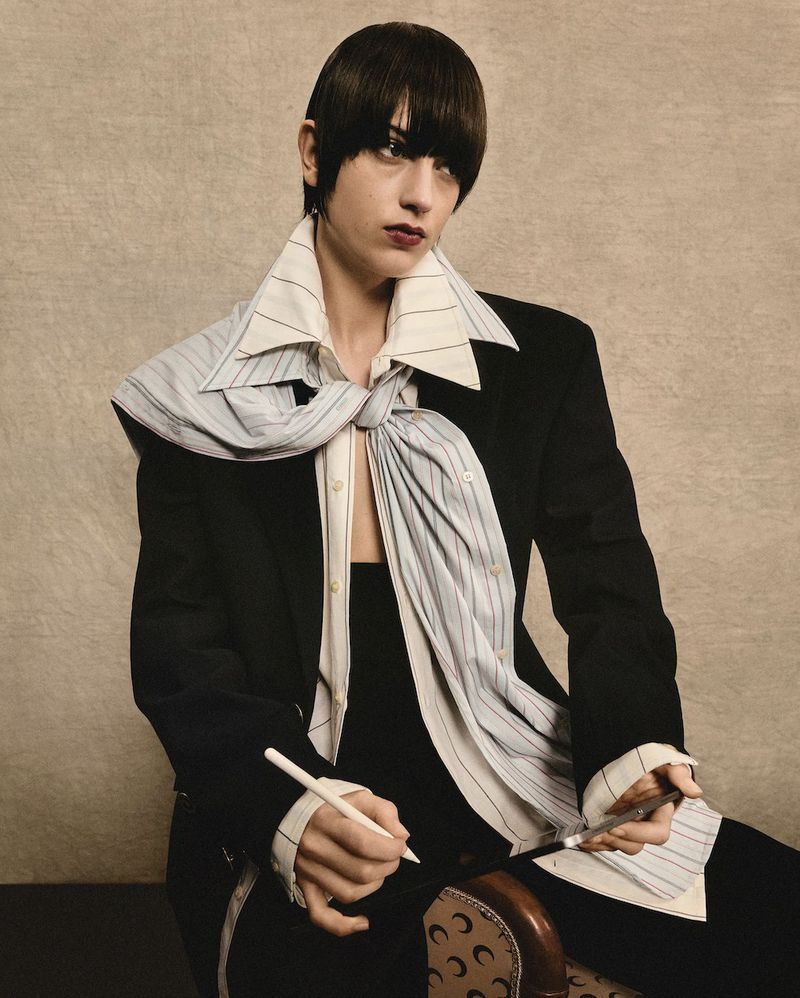 A woman with dark hair in a black blazer and a large striped tie is posing with pens in her hands in a neutral indoor setting.